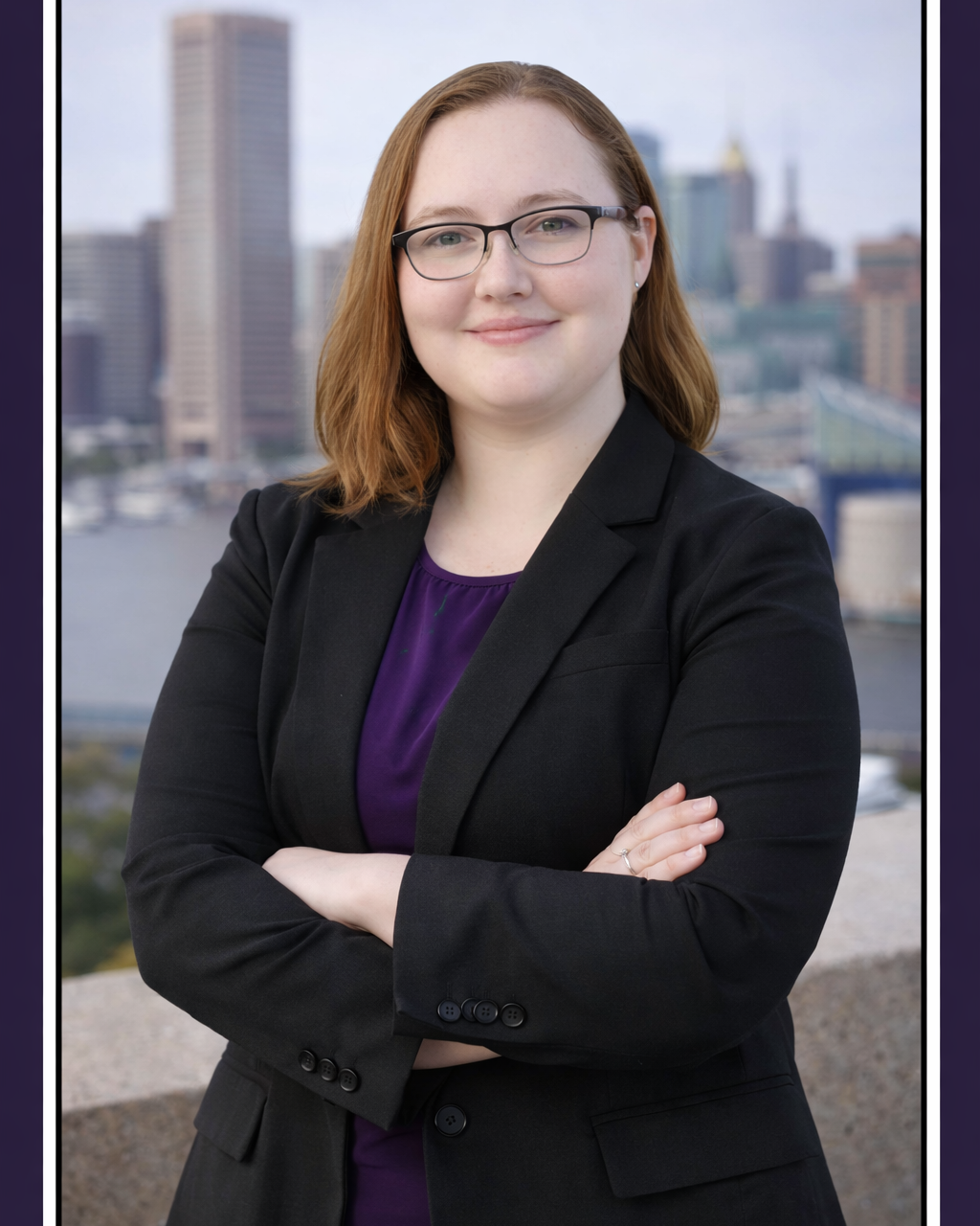 Businesswoman with glasses and red hair standing outdoors with a cityscape background, arms crossed, wearing a black blazer and purple blouse.