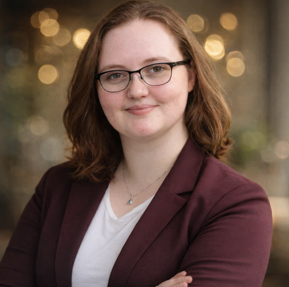 A young woman with shoulder-length auburn hair, wearing glasses, a maroon blazer, and a white top, smiling at the camera with a blurred bokeh background.